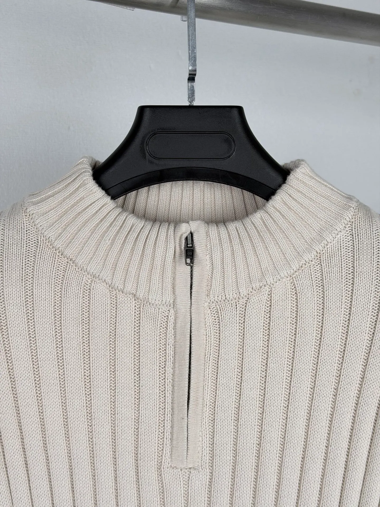 BALENCIAGA/Balenciaga Cola Wave Half-Zip Pit Knitted Sweater Ready-made garments are made of longitudinal pit texture knitted gray fabric. The weaving stage has high standards for yarn tension and machine yarn feeding speed. The width of the vertical grooves and the concave and convex texture of the entire body of the garment must be highly uniform. The production process is prone to defects such as texture deviation and density gaps, and the gray fabric screening loss is large. After the gray fabric is off the machine, it is pre-shrunk and heat-set to release the potential shrinkage of the fabric in advance and prevent the subsequent distortion and size deformation of the finished garments in the washing water. The body of the garment is equipped with a half-zip stand-up collar structure. The collar ribs and body pit stripes require vertical alignment, and the splicing process requires precise alignment of the texture direction. The zipper welt uses a blind-stitching process to ensure smooth opening and closing of the zipper and avoid local vertical grain stretch deformation caused by sewing pull. Sewing workers need to continuously control the fabric feeding tension to balance the elasticity of the fabric and the stability of the threading. Drop-shoulder silhouette pieces are cut strictly along the pitted texture to prevent diagonal cuts from damaging the fabric texture. The printing of the wave monogram on the back is the core technical difficulty of this model. The surface of the concave and convex pit fabric is undulating and cannot be printed directly like flat fabrics. During production, the tension of the printing area is first fixed and leveled, and the layered alignment offset printing process is used to complete the printing of wave graphics and letter logos in sequence. Multi-layer pattern overprinting requires extremely high precision, and it is easy to produce ghosting and misalignment defects. The ink ratio has been adjusted to take into account the fullness of the pattern and ensure that the printing is not prone to cracking and falling off when the fabric is stretched. The cuffs and hem ribs are woven from pitted fabrics of the same system, and the elastic density matches the body parameters to avoid problems with elasticity imbalance and wavy edges after wearing. After the garments are sewn, detailed inspection is carried out to screen for sewing defects and printing defects. Finally, the low-temperature wet finishing process is used to uniformly correct the pattern and texture flatness. From knitting and weaving, precision cutting, structural sewing, multi-layer overprinting to garment styling, the process chain is complicated. Failure to control any process standard will directly affect the overall look and feel of the garment. Size:XS S M L